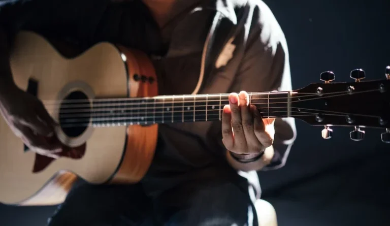 Close de uma pessoa tocando violão, focando nas mãos no braço do instrumento, iluminadas por uma luz suave ao fundo.